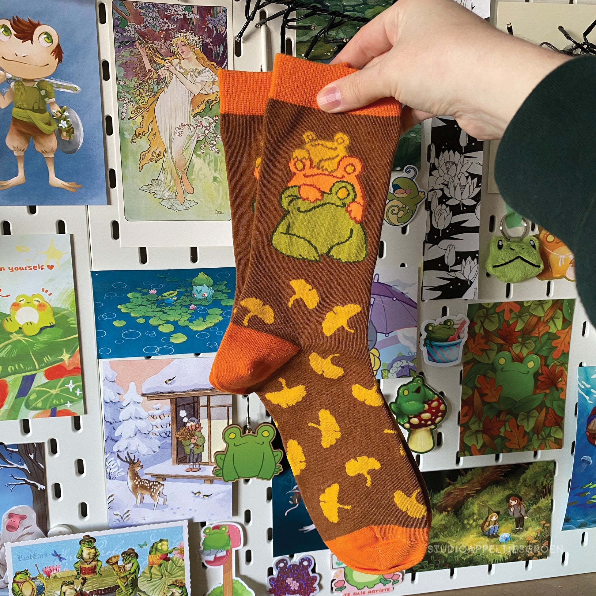 Orange with brown socks depicting three frogs stacked on top of each other, with a yellow ginkgo leaves pattern, held up by a hand wearing a green sleeve.