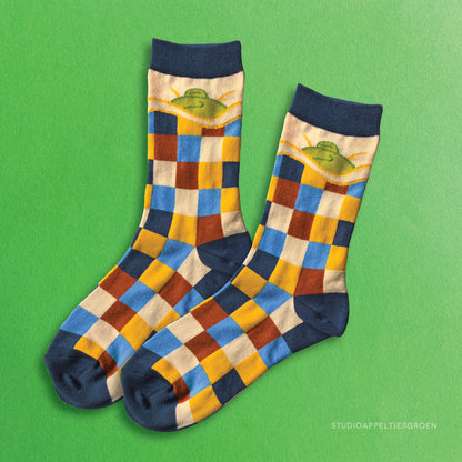 Frog socks depicting a sleepy frog underneath a blanket. The blanket being the checkered pattern of blue, dark blue, yellow, cream and a brown red.