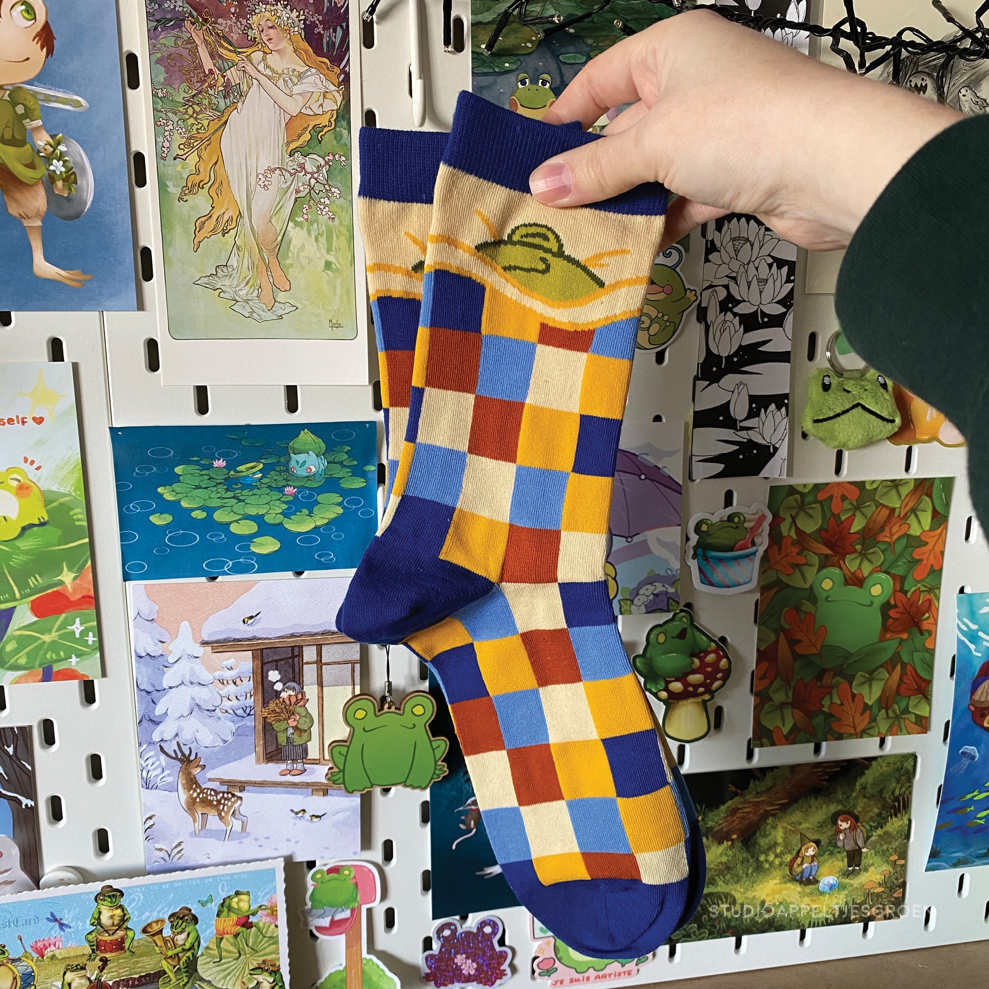 Frog socks depicting a sleepy frog underneath a blanket. The blanket being the checkered pattern of blue, dark blue, yellow, cream and a brown red. The socks are being held up by a hand wearing a green sleeve.