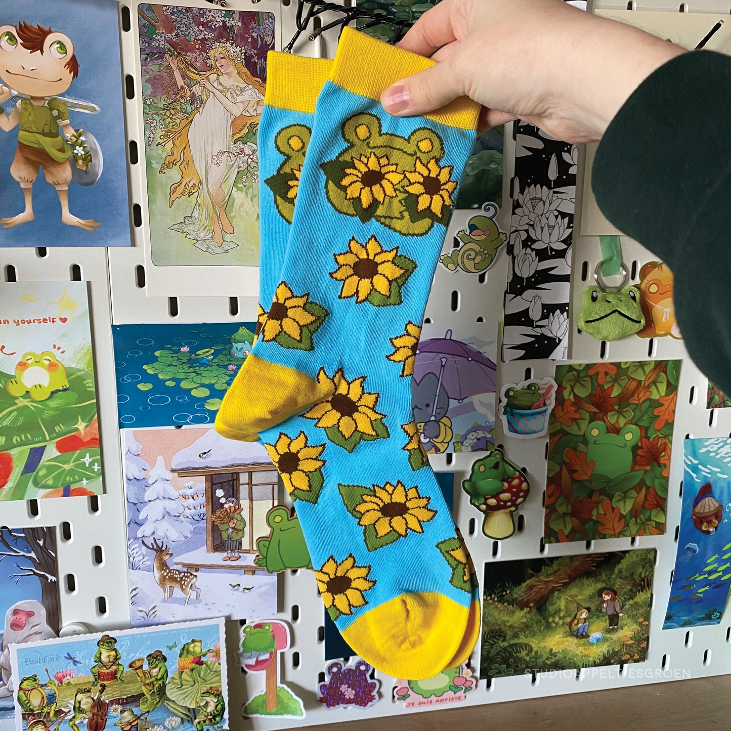 Frog socks in bright blue and yellow depicting a frog holding up two sunflowers and a pattern of other sunflowers on the rest of the sock. The socks are being held up by a hand wearing a green sleeve.