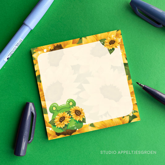 Notebook with sunflower and frog design on a green surface with stationery items.